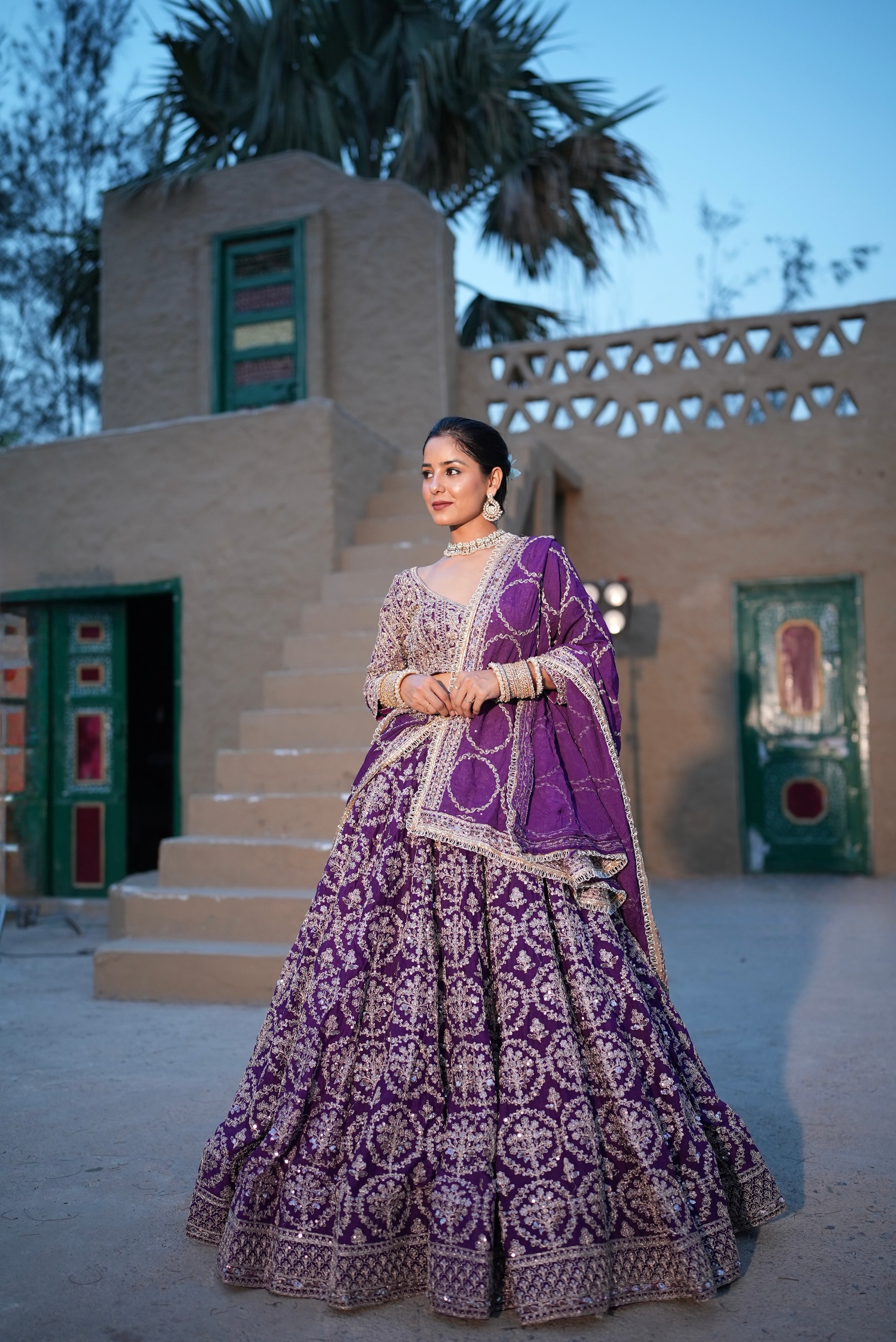 Woman in a purple traditional outfit standing in front of a building with green doors.