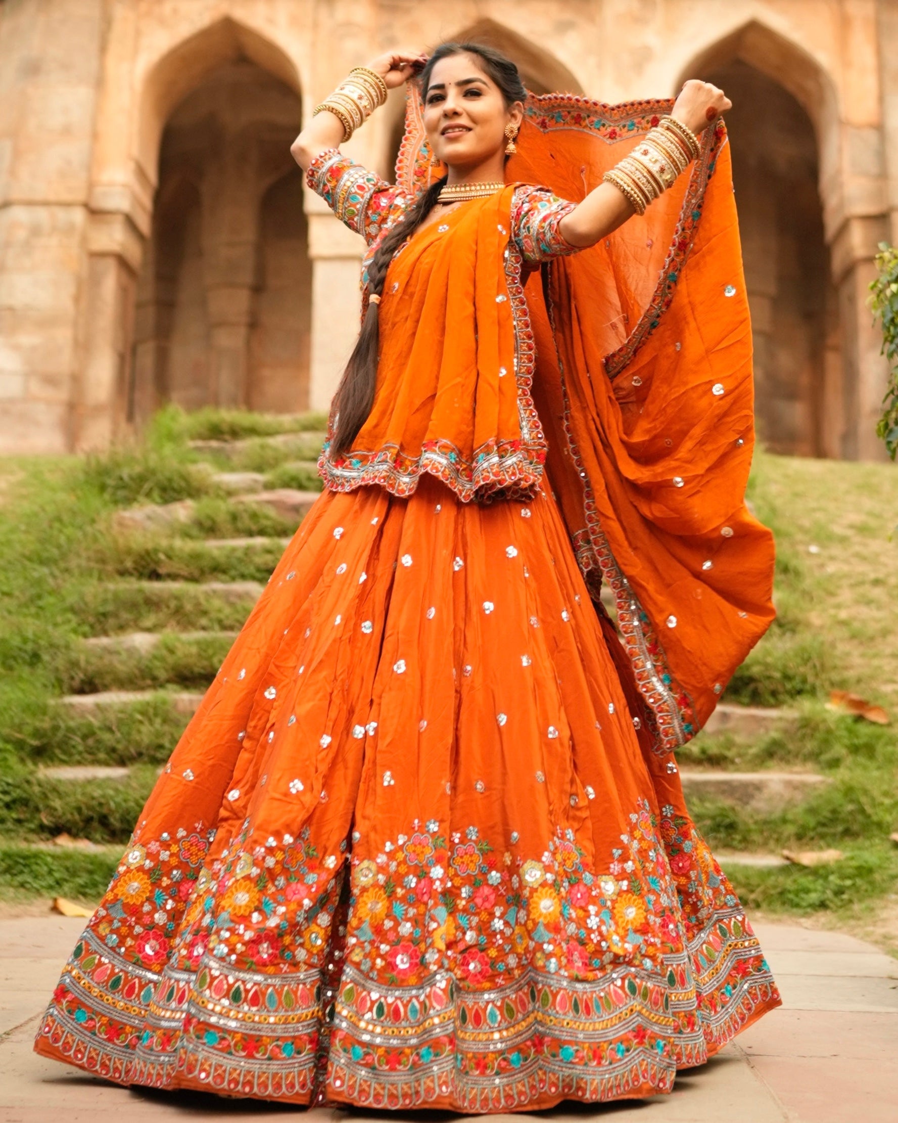 Woman in an orange traditional outfit with floral patterns standing outdoors.