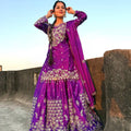 Woman in a purple traditional outfit with intricate designs, standing against a clear sky.